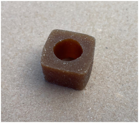 A brown, cube-shaped piece of dog treat with a circular hole in the center, resting on a textured beige surface.