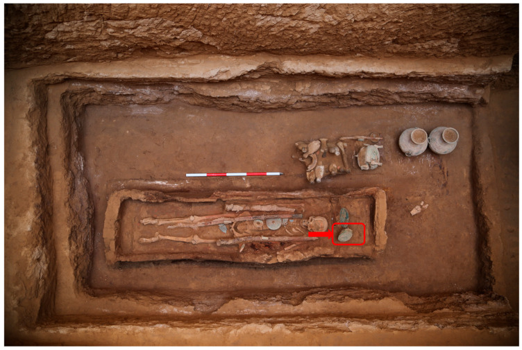 The excavation site of the yellow-greenish fiber sheet on the bronze mirror (the excavation location of the mirror was indicated by the red box and arrow).